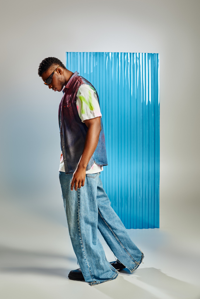 Вид збоку модної афроамериканської моделі в сонцезахисних окулярах, джинсовий жилет і джинси, що ходять на сірому з синім полікарбонатним листом на фоні, стійка мода, одяг для саморобки
 - Фото, зображення