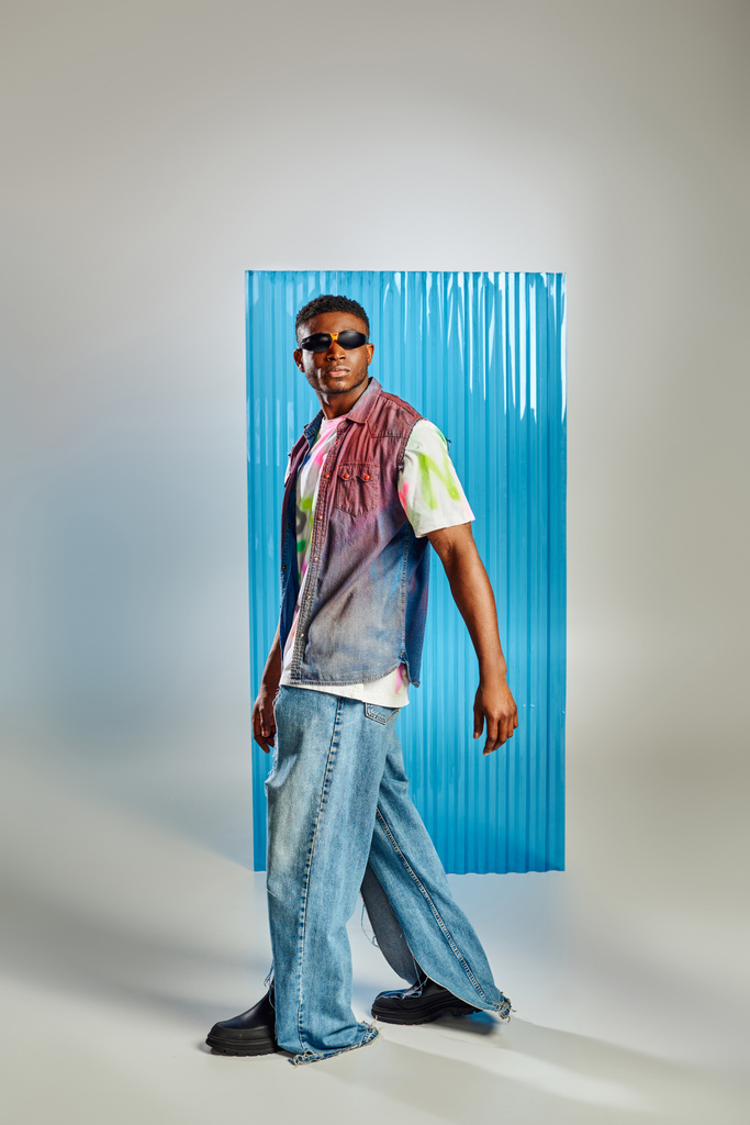Modny młody model afroamerykański w dżinsowej kamizelce, kolorowy t-shirt i podarte dżinsy spacerujące na szarym tle z niebieskim prześcieradłem poliwęglanowym, zrównoważona moda, odzież DIY - Zdjęcie, obraz