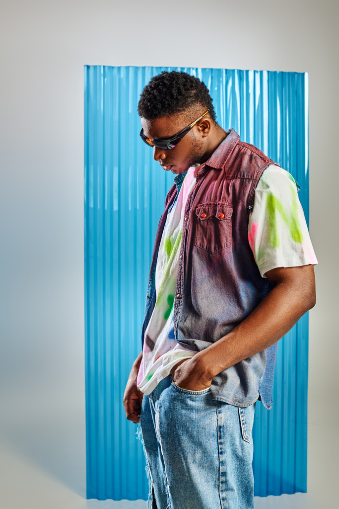 Trendy junger afroamerikanischer Mann mit Sonnenbrille, buntem T-Shirt und Jeansweste, die Hand in der Hosentasche einer zerrissenen Jeans auf grau mit blauem Polycarbonat-Laken im Hintergrund, DIY-Kleidung - Foto, Bild