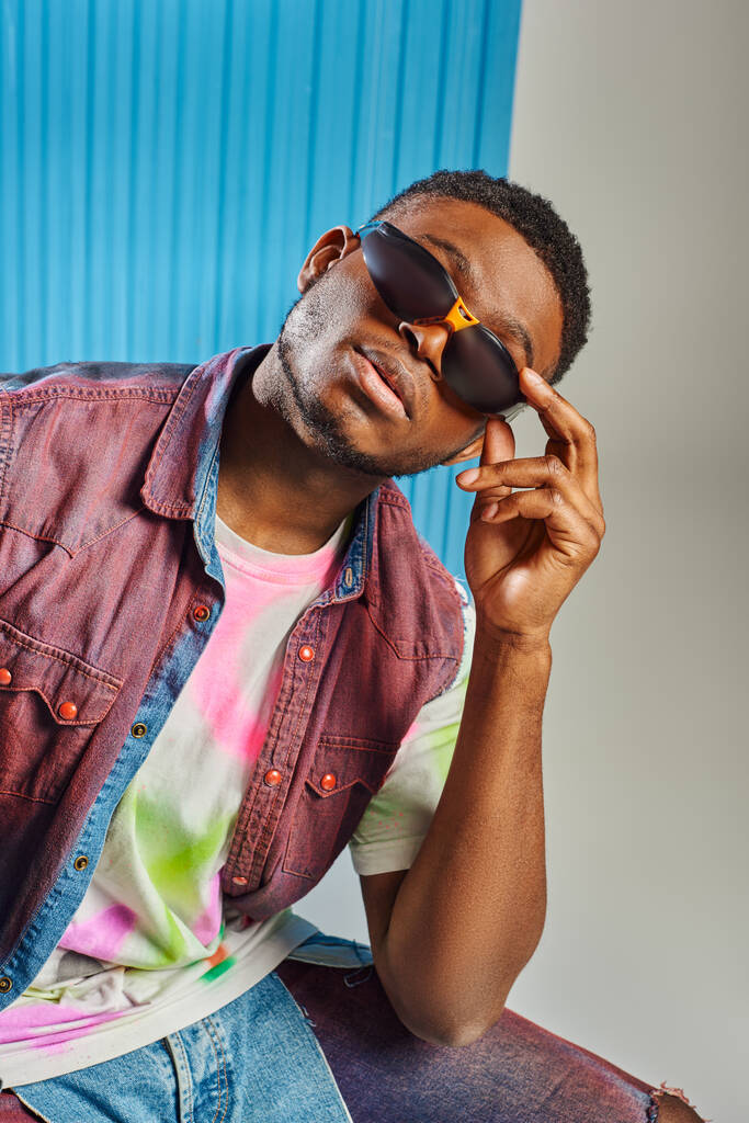 Modelo afroamericano jovem na moda em colete de ganga e camiseta colorida tocando óculos de sol em cinza com folha de policarbonato azul no fundo, tiro de moda, roupas DIY, estilo de vida sustentável  - Foto, Imagem
