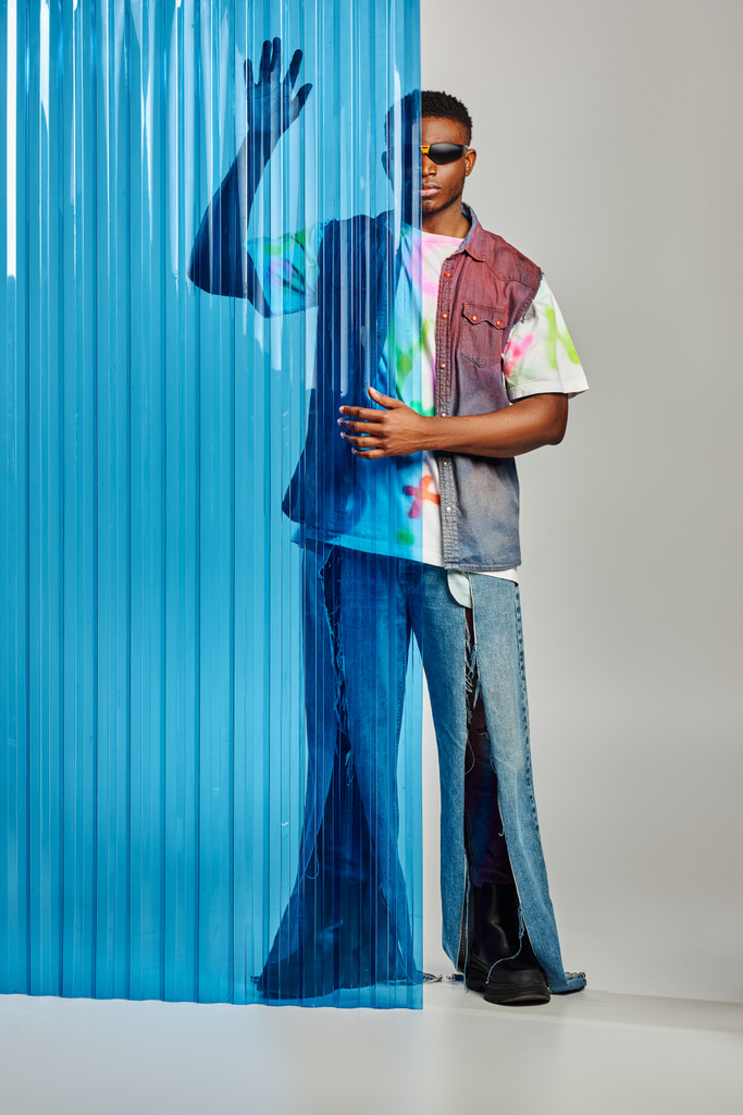 Afroamerikaner mit Sonnenbrille, zerrissenen Jeans und bunter Jeansweste posiert neben blauem Polycarbonat-Laken und steht auf grauem Hintergrund, Mode-Shooting, DIY-Kleidung, nachhaltiger Lebensstil  - Foto, Bild