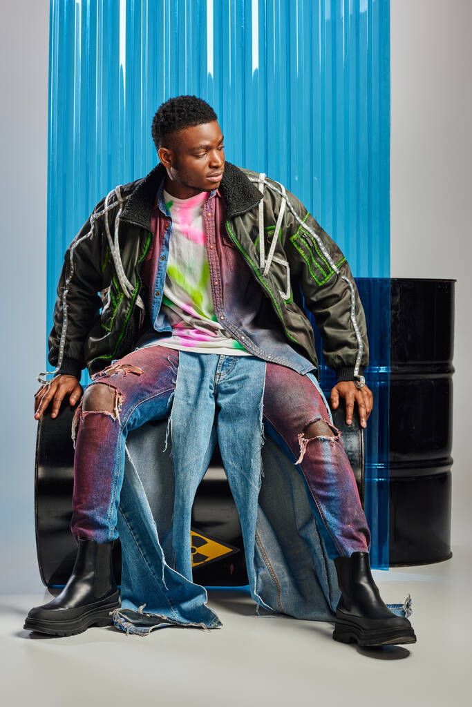 Stilvoller afroamerikanischer Mann in Outwear-Jacke mit Bleistreifen und bunt zerrissenen Jeans sitzt auf einem Benzinfass neben blauem Polycarbonat-Laken und auf grauem Hintergrund, DIY-Kleidung, nachhaltiger Lebensstil  - Foto, Bild