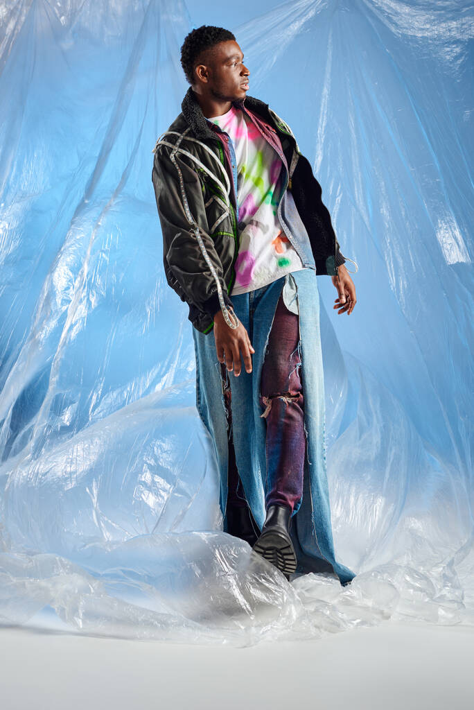 Modieuze Afro-Amerikaanse man in outwear jas met led strepen en gescheurde jeans lopend op glanzend cellofaan op blauwe achtergrond, urban outfit en moderne pose, DIY kleding, duurzame levensstijl  - Foto, afbeelding