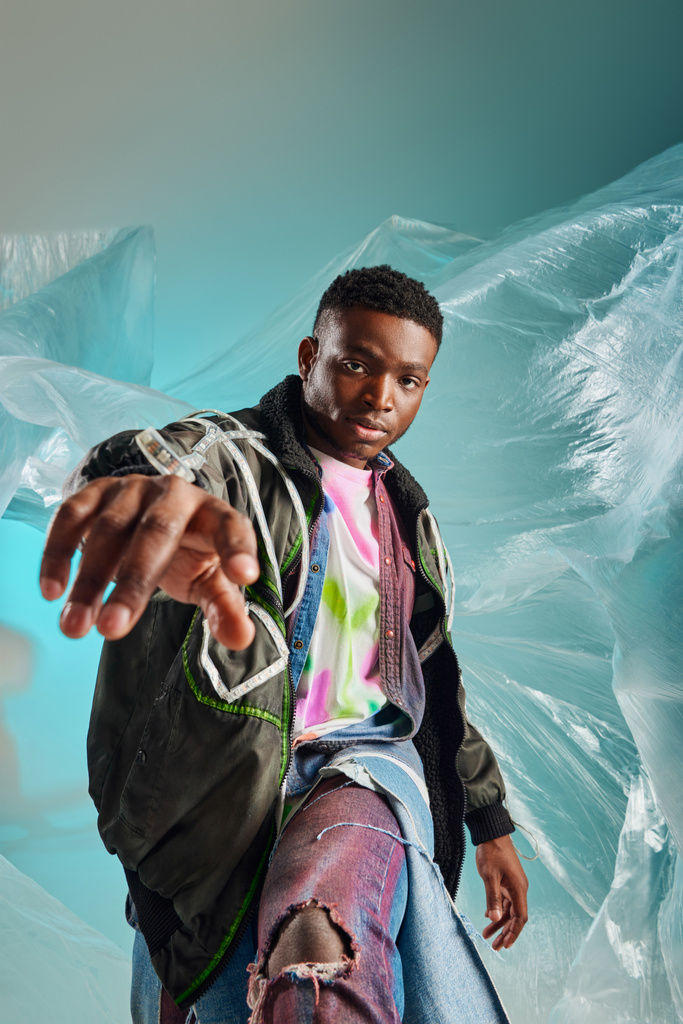 Portret van een zelfverzekerde jonge Afro-Amerikaanse man in outtwear jas en gescheurde jeans poseren met cellofaan op turquoise achtergrond, stedelijke outfit en moderne pose, creatieve expressie, DIY kleding  - Foto, afbeelding