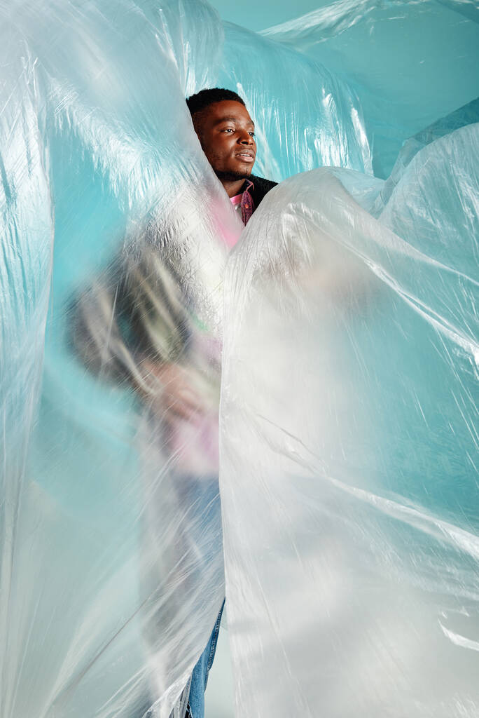 Young african american man with trendy hairstyle looking away while standing in cellophane on turquoise background, urban outfit and modern pose, creative expression, DIY clothing  - Photo, Image