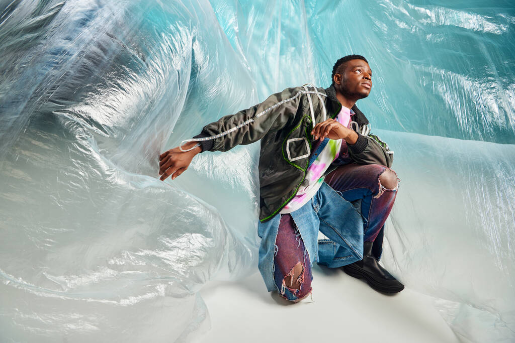Nuori afroamerikkalainen malli päällystakissa, jossa on led-raidat ja revityt farkut, jotka katsovat pois lähellä sellofaania turkoosilla taustalla, kaupunkiasussa ja modernissa asennossa, luova ilme, DIY-vaatteet  - Valokuva, kuva