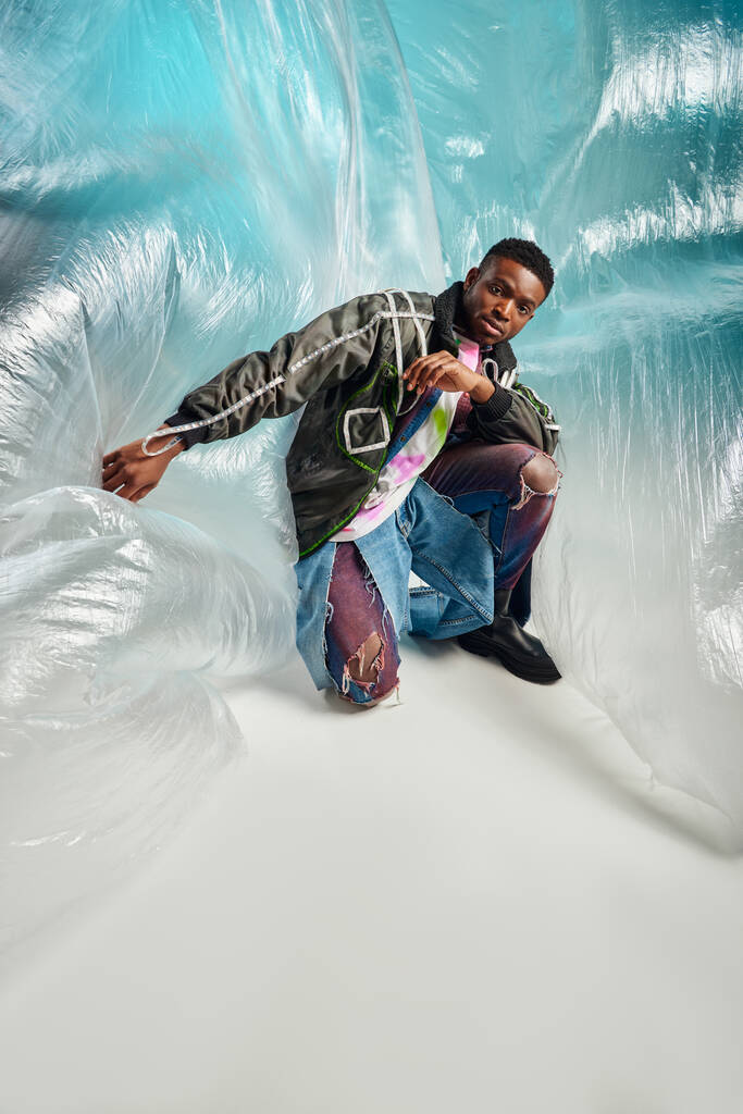 Modelo americano africano jovem elegante e confiante em jaqueta de roupa interior e jeans rasgados olhando para a câmera perto de celofane em fundo turquesa, expressão criativa, roupas DIY  - Foto, Imagem