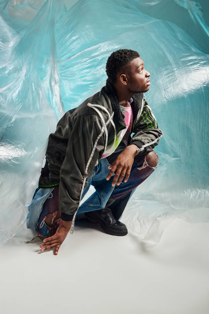 Vista lateral do homem afroamericano bem parecido em jaqueta de roupas com listras led e jeans rasgados olhando para cima perto de celofane brilhante em fundo turquesa, expressão criativa, roupas DIY  - Foto, Imagem