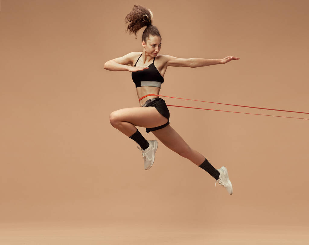 Dynamic image of sportive, fit, strong young girl training, in motion, jumping with fitness string against light brown studio background. Sportive lifestyle, beauty, body care, fitness, health concept - Photo, Image