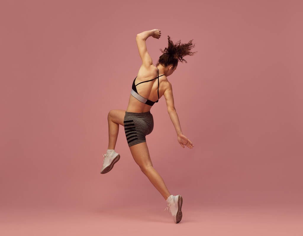 Cvičení celého těla. Dynamický obraz mladé sportovní ženy v tréninku sportovního oblečení, pózující v pohybu na růžovém pozadí studia. Koncept sportovního životního stylu, krásy, péče o tělo, fitness, zdraví - Fotografie, Obrázek