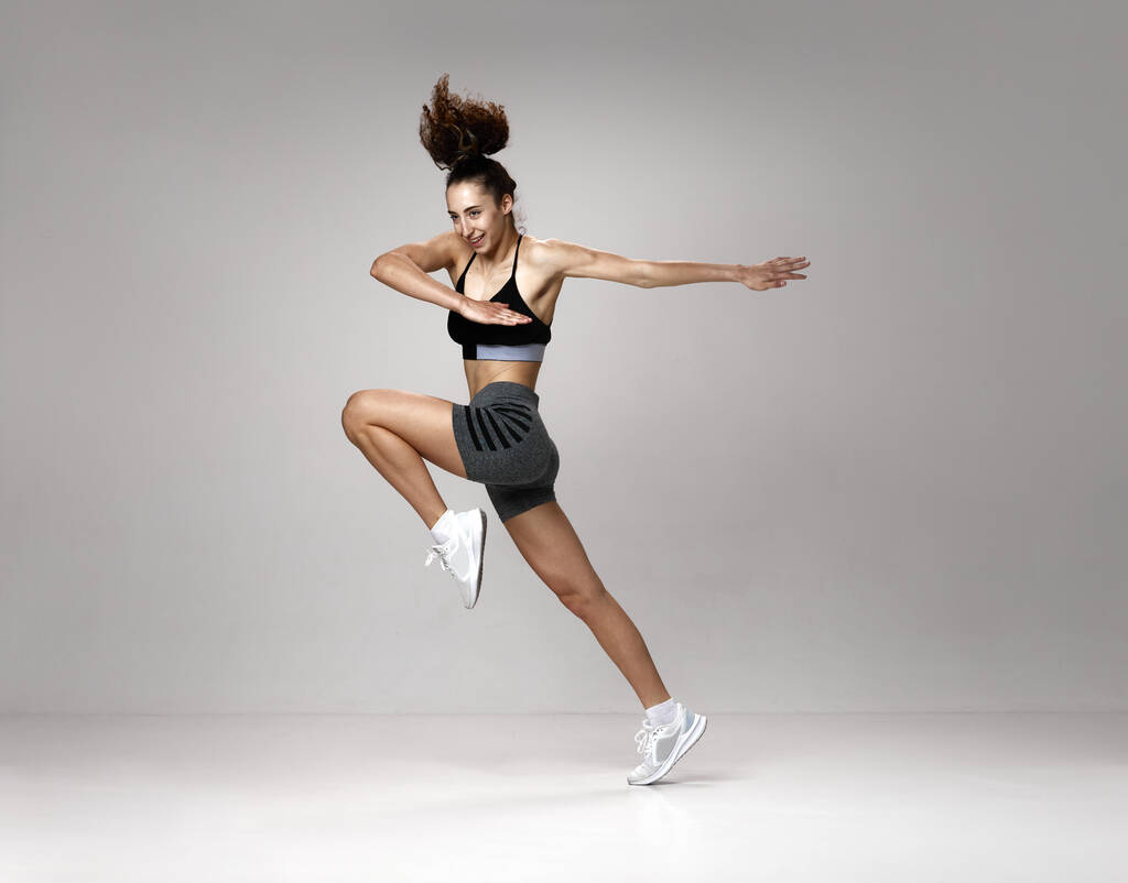 Hoog springen. Dynamisch beeld van jonge mooie vrouw in sportkleding training, poseren in beweging tegen grijze studio achtergrond. Concept van sportieve levensstijl, schoonheid, lichaamsverzorging, fitness, gezondheid - Foto, afbeelding