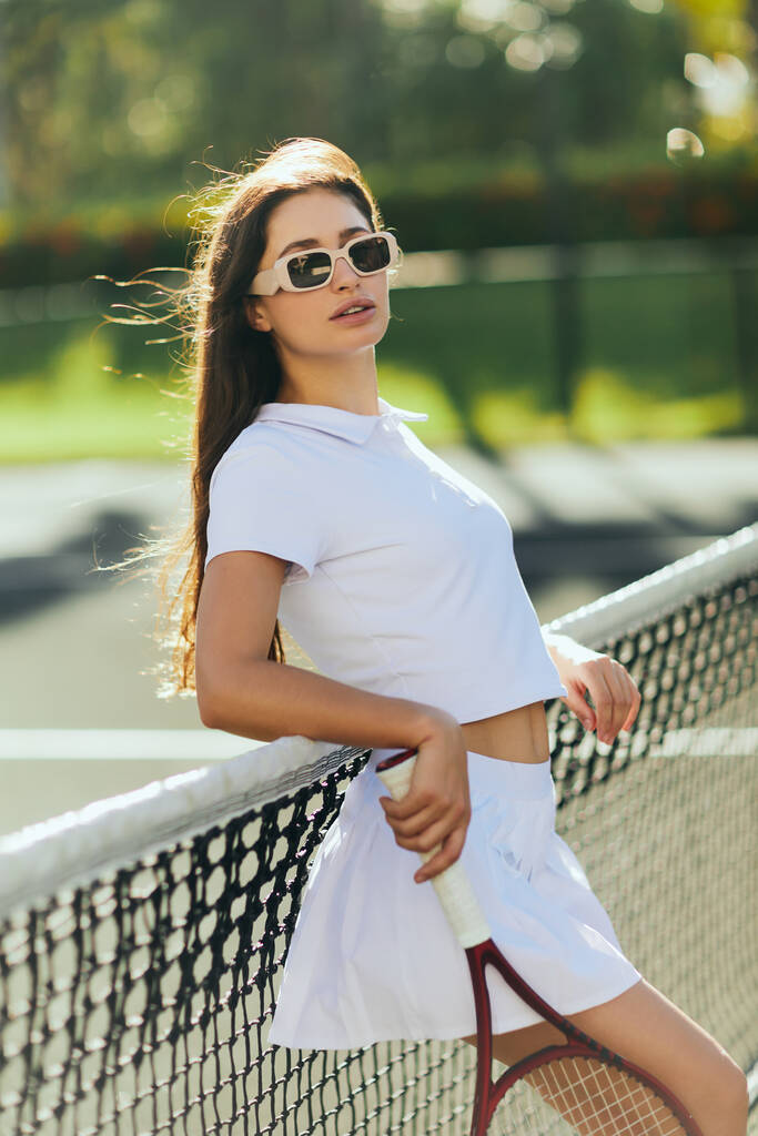 tennis court in Miami, sporty young woman with brunette long hair standing in white outfit and sunglasses while holding racket near tennis net, blurred background, iconic city, looking at camera  - Photo, Image