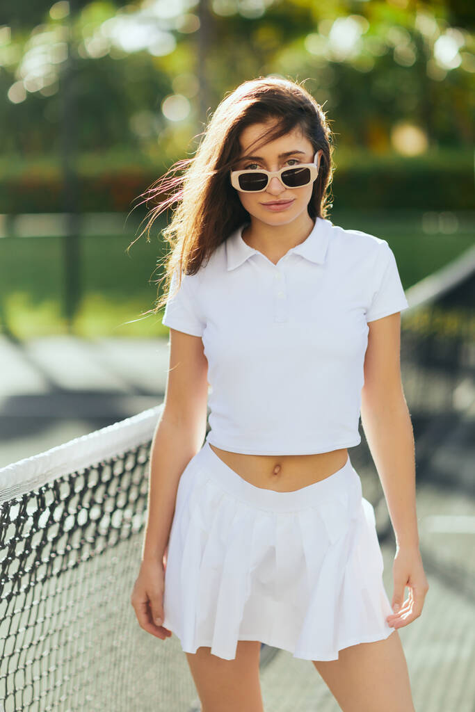 portrait of stylish young woman with brunette long hair standing in white outfit and sunglasses near tennis net, blurred background, wind, tennis court in Miami, iconic city, Florida, sunny day  - Photo, Image