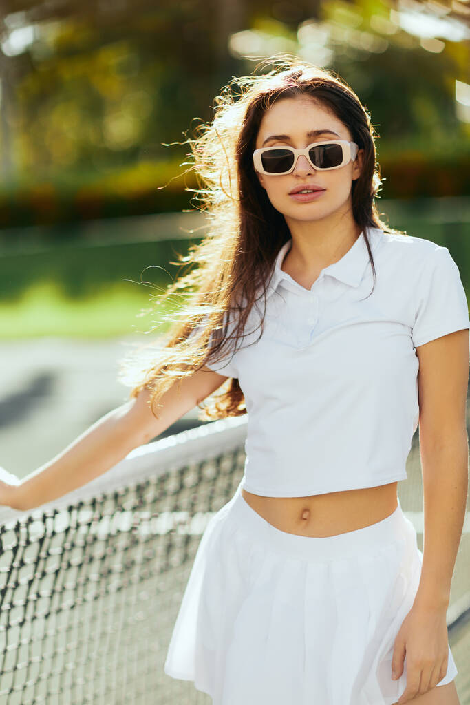 portrait of pretty young woman with brunette long hair standing in white outfit and sunglasses near tennis net, blurred background, tennis court in Miami, Florida, iconic city, sunny day  - Photo, Image