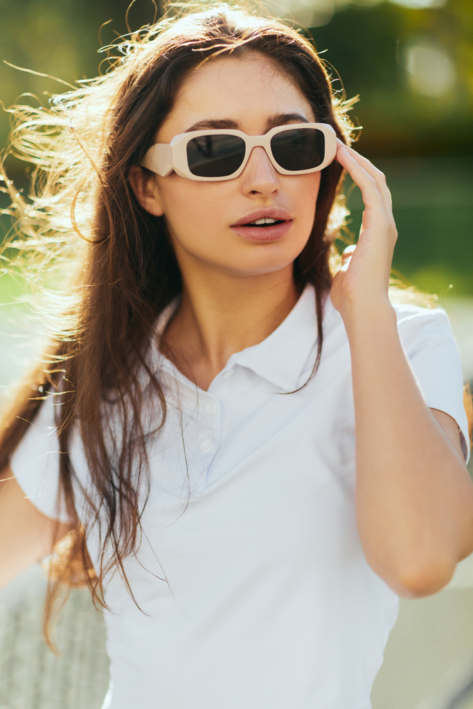 πορτρέτο της κομψής νεαρής γυναίκας με καστανά μακριά μαλλιά στέκεται σε λευκό ντύσιμο και γυαλιά ηλίου κοντά θολή δίχτυ τένις στο παρασκήνιο, γήπεδο τένις στο Μαϊάμι, Φλόριντα, εικονική πόλη, ηλιόλουστη μέρα  - Φωτογραφία, εικόνα