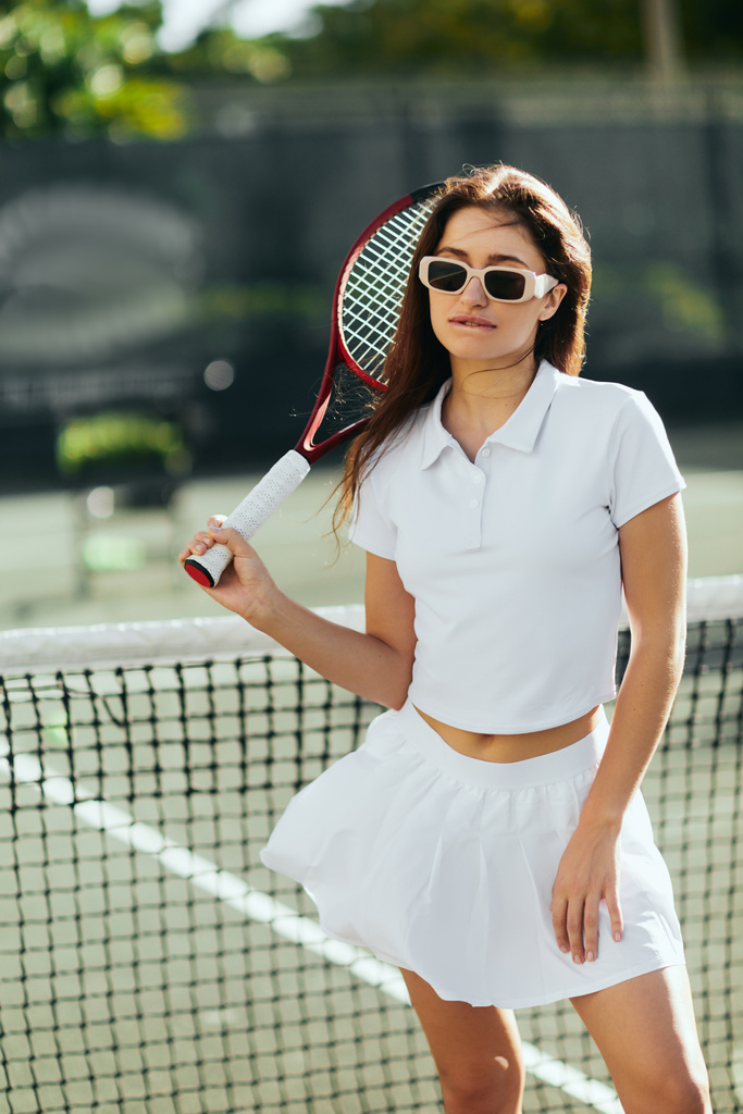 actieve levensstijl, brunette en jonge vrouw staan in stijlvolle outfit en zonnebril terwijl het houden van racket in de buurt van tennis net, wazig achtergrond, iconische stad, Miami, Florida, zonnige dag, zomer  - Foto, afbeelding