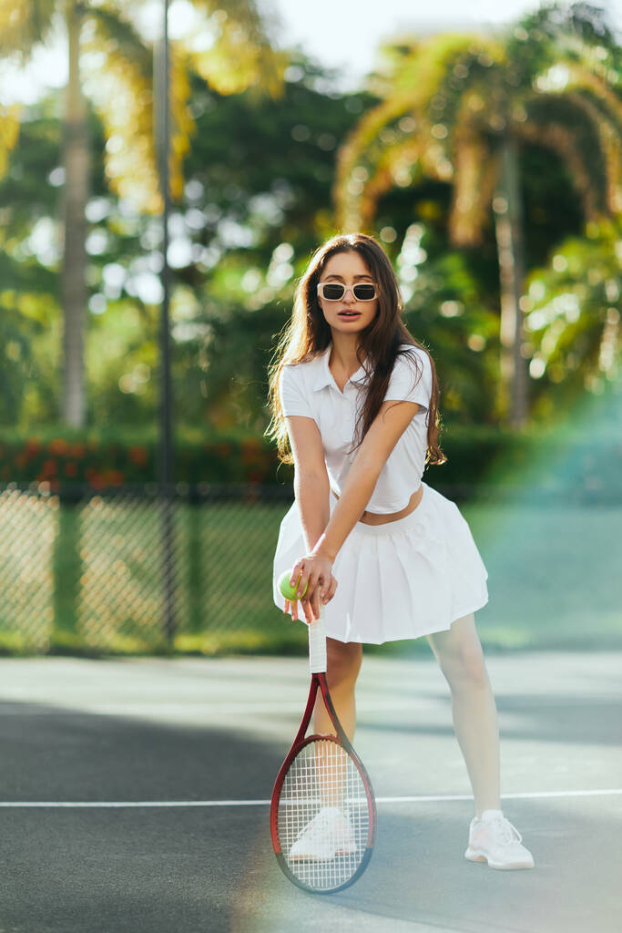 энергичная брюнетка с длинными волосами, стоящая в стильном белом наряде и держащая ракетку с мячом на теннисном корте в Майами, Флорида, солнечный день, пальмы на размытом фоне, теннисная юбка   - Фото, изображение