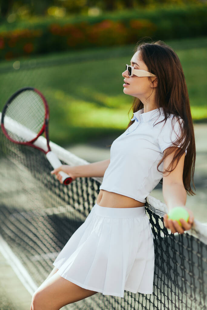 Tennisplatz in Miami, athletische junge Frau mit langen Haaren in weißem Outfit und Sonnenbrille, während sie verschwommenen Schläger und Ball in der Hand hält und sich an Tennisnetz lehnt, grüner Hintergrund, ikonische Stadt  - Foto, Bild