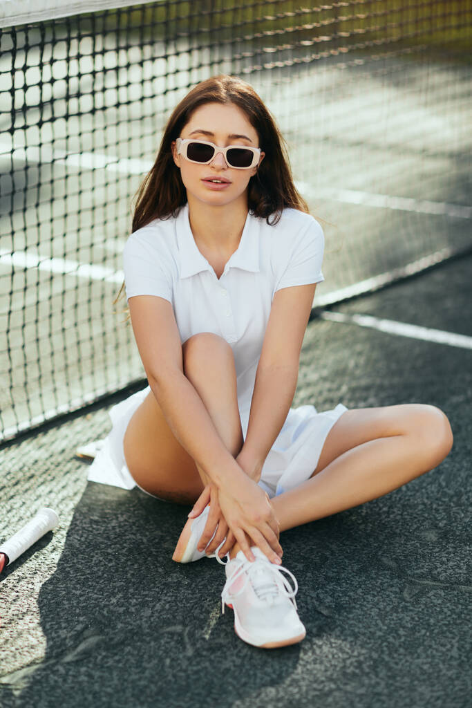 teniszpálya Miami, sportos fiatal nő barna hosszú haj ül fehér ruhában, cipő és napszemüveg közelében tenisz háló, homályos háttér, ikonikus város, fizikai aktivitás  - Fotó, kép