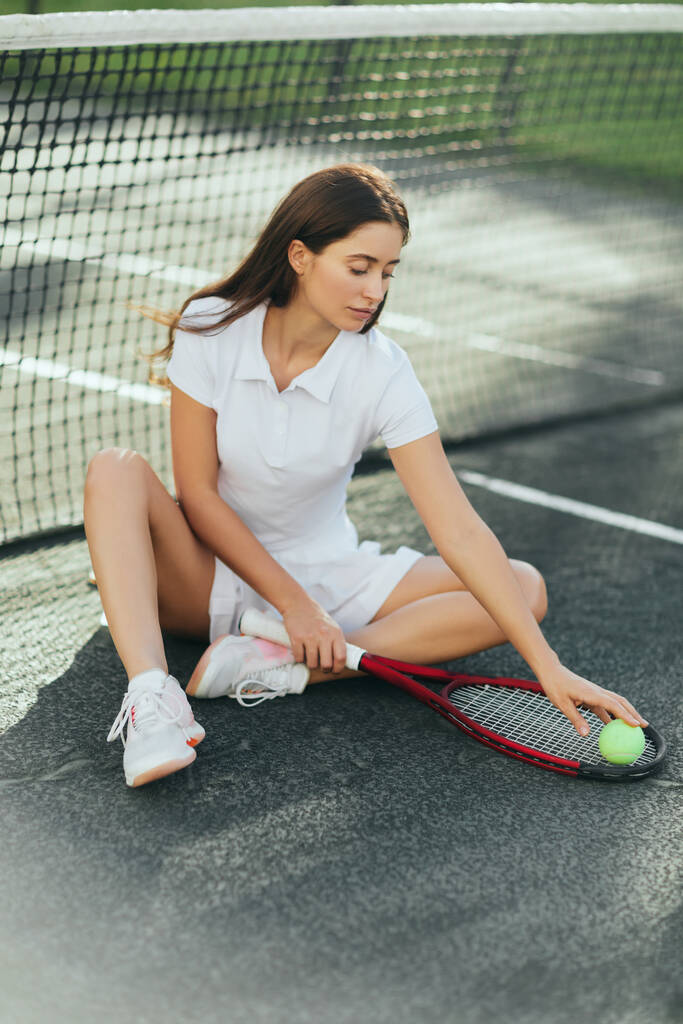γυναίκα τενίστρια ξεκουράζεται μετά το παιχνίδι, νεαρή γυναίκα με μακριά μαλλιά κάθεται σε λευκό ντύσιμο και κρατώντας ρακέτα με μπάλα κοντά στο δίχτυ του τένις, θολή φόντο, Μαϊάμι, εικονική πόλη, γήπεδο τένις  - Φωτογραφία, εικόνα