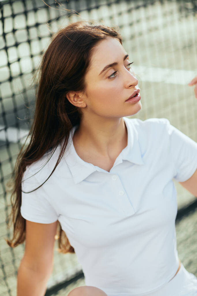 портрет привлекательной молодой женщины с длинными волосами брюнетки в белой рубашке поло и отводящей взгляд после тренировки на теннисном корте, теннисная сетка на размытом фоне, Майами, Флорида  - Фото, изображение