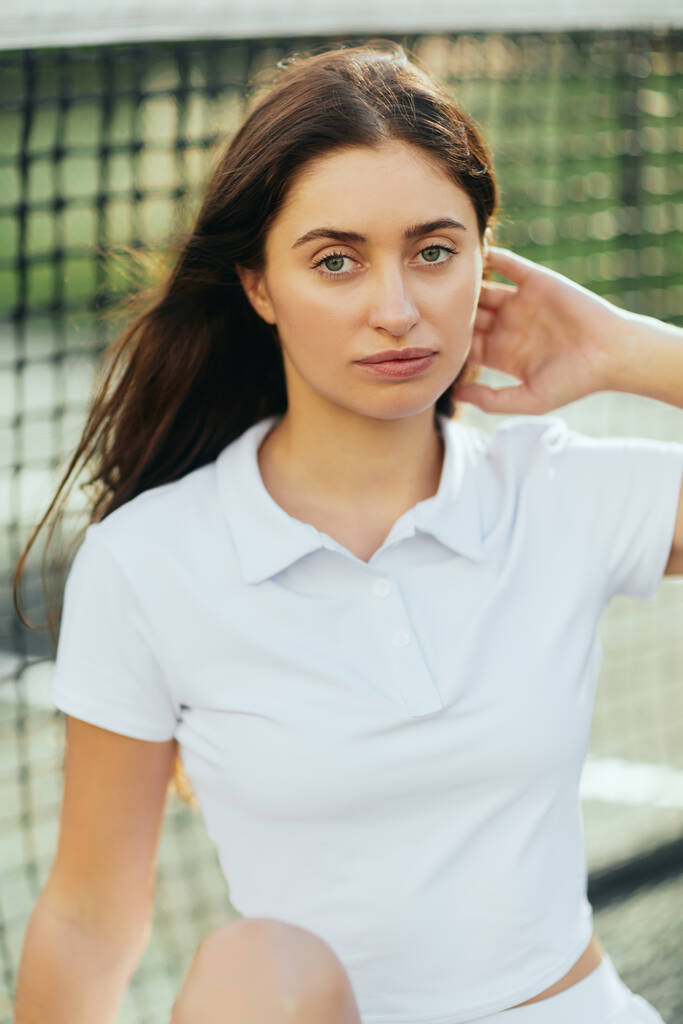 portret van mooie jonge vrouw met lang brunette haar in wit poloshirt en kijkend naar camera na training op tennisbaan, tennisnet op wazige achtergrond, Miami, Florida  - Foto, afbeelding