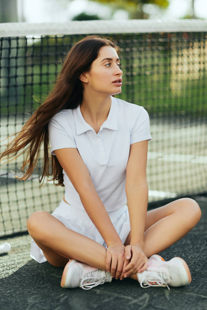 distraído jugador femenino en la cancha de tenis, mujer joven con el pelo largo sentado con las piernas cruzadas en traje blanco y zapatillas de deporte y mirando lejos cerca de la red de tenis, fondo borroso, Miami, tiempo de inactividad - Foto, imagen