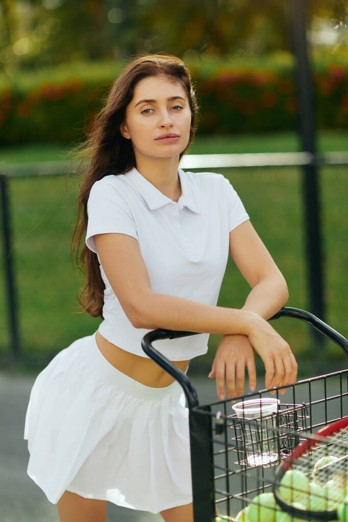 здоровые привычки, спортивная молодая женщина с брюнеткой волосы стоят в белом наряде с юбкой и рубашкой поло и опираясь на тележку с мячами и ракеткой, размытый фон, теннисный корт в Майами  - Фото, изображение