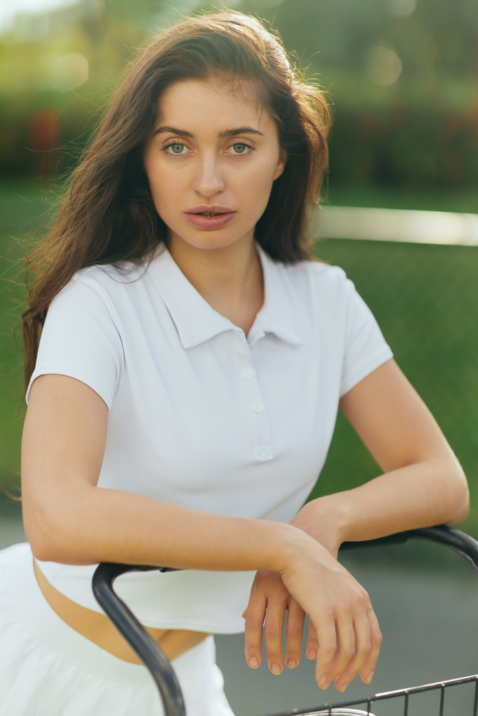 pretty tennis player, sporty young woman with brunette hair standing in white polo shirt near tennis cart, blurred green background, looking at camera, tennis court in Miami  - Photo, Image