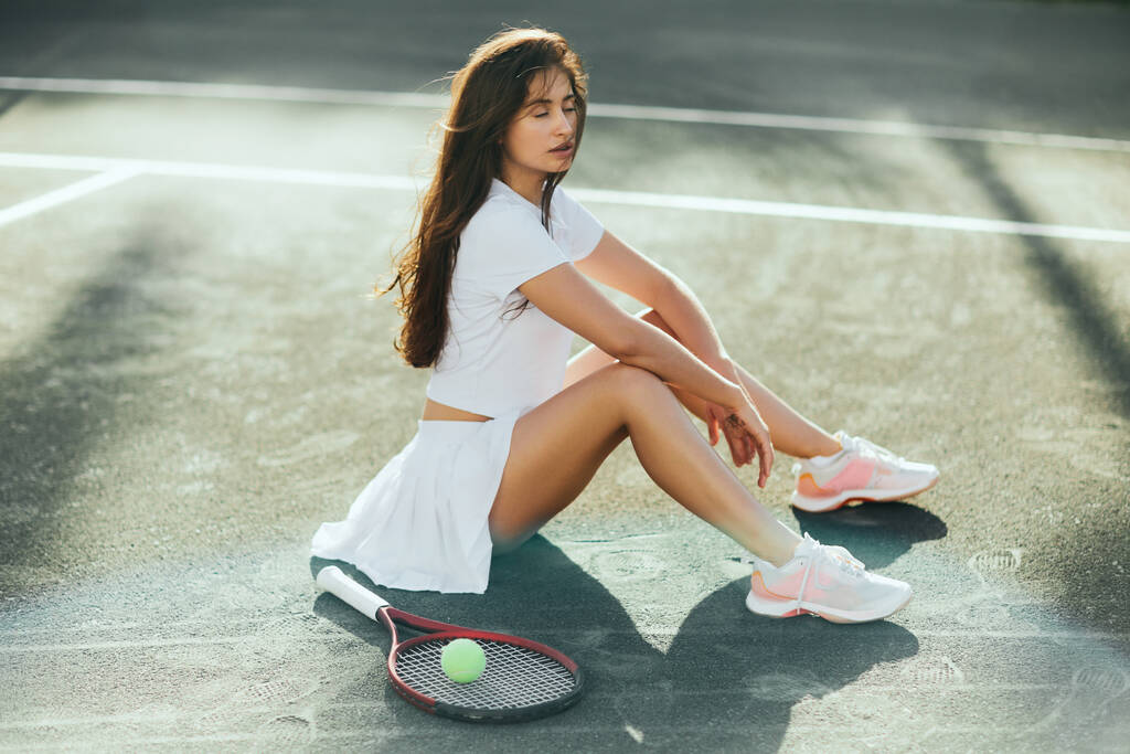hráčka odpočívající po hře, brunetka mladá žena se zavřenýma očima sedící v bílém kostýmu v blízkosti rakety s míčem na asfaltu, Miami, tenisový kurt, prostoje, stíny, slunečný den - Fotografie, Obrázek