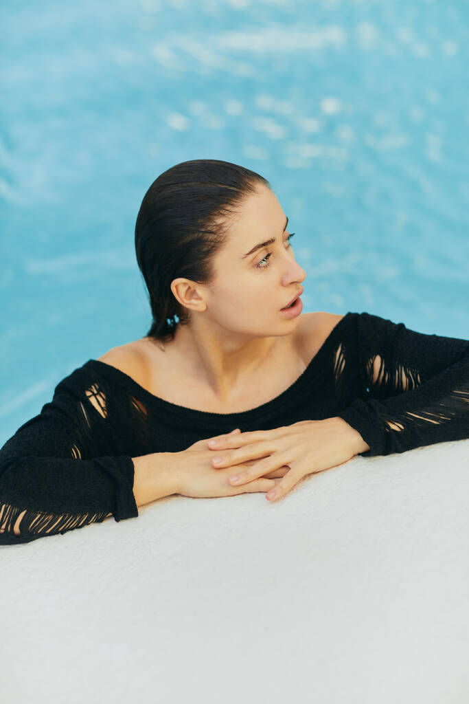 Mooie jonge vrouw met gebruinde huid in zwarte badmode in openbaar zwembad, poseren en genieten van haar zomervakantie in luxe resort in Miami, geen make-up look - Foto, afbeelding