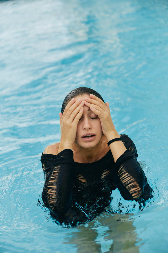 роскошный курорт в Майами, красивая женщина с тонированной кожей, касающаяся своего мокрого лица после купания в общественном бассейне, позируя и наслаждаясь своим летним отпуском, без макияжа  - Фото, изображение