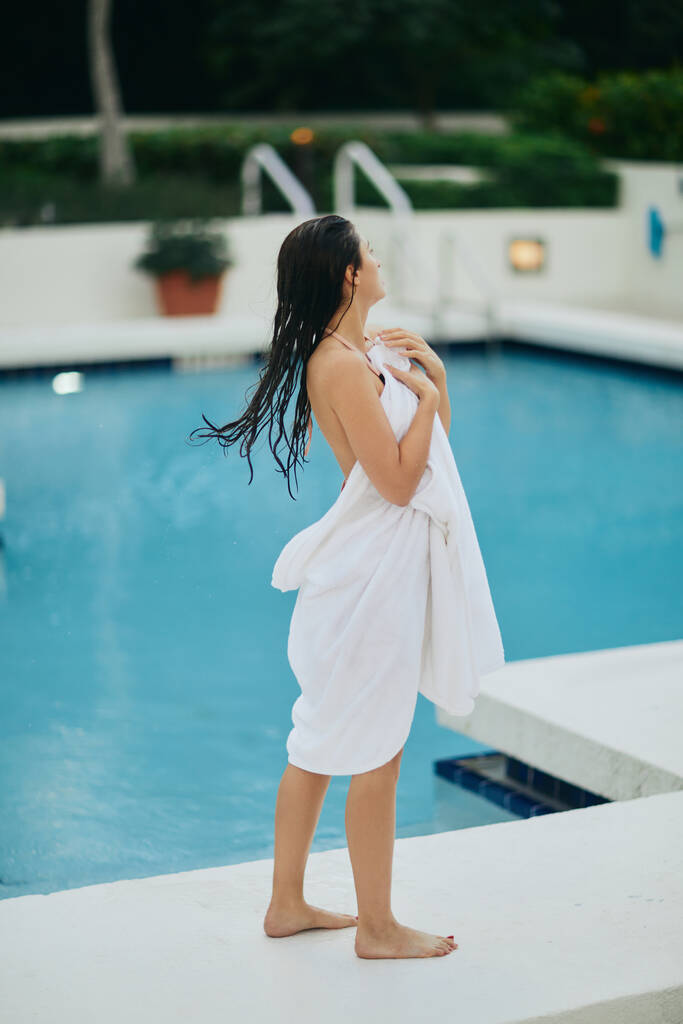 jeune femme brune aux cheveux mouillés enveloppée dans une serviette blanche debout à côté de la piscine extérieure avec de l'eau chatoyante à Miami, escapade estivale, jeunesse, détente au bord de la piscine, mode vacances  - Photo, image