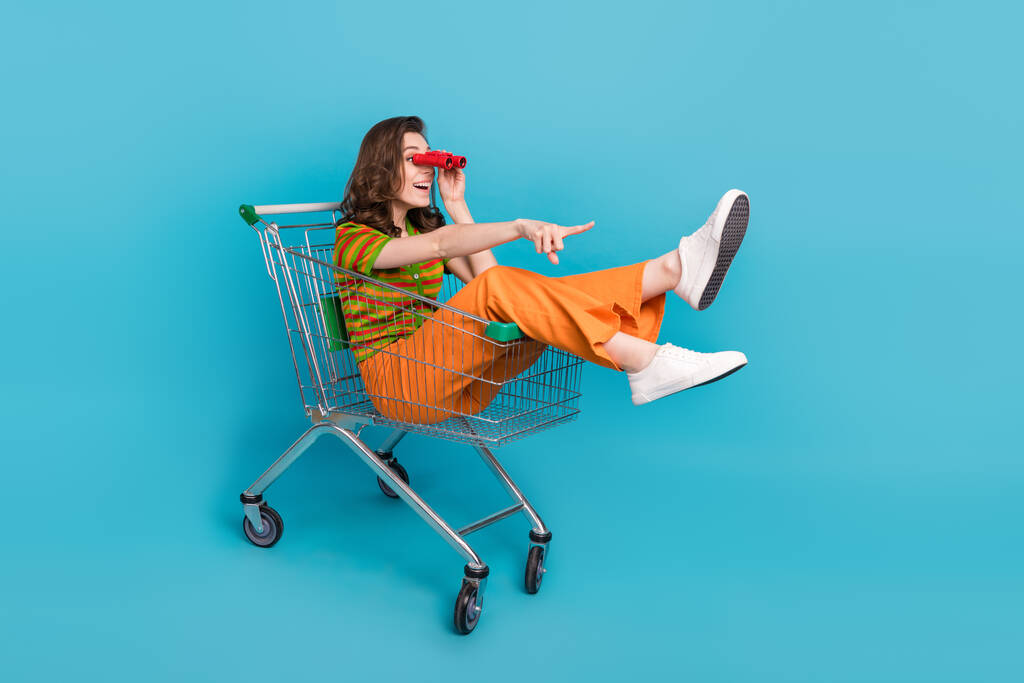 Полноразмерная фотография экстатической симпатичной женщины, сидящей в картонке для покупок, смотрит в бинокль, направляя пустое пространство, изолированное на синем цветовом фоне. - Фото, изображение