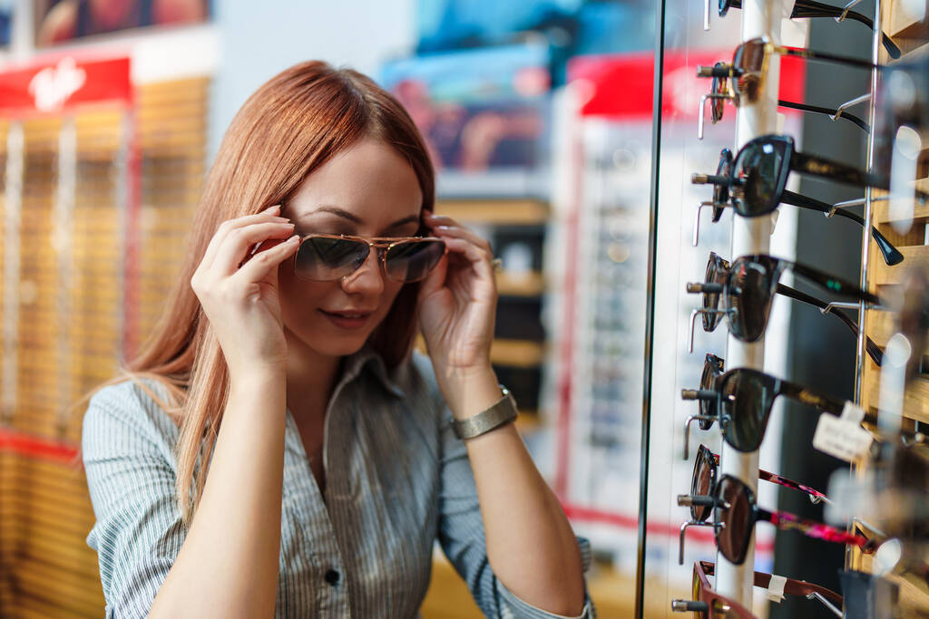 Beautiful and fashionable redhead woman choosing eyeglasses frame in a modern optical store. - Photo, Image
