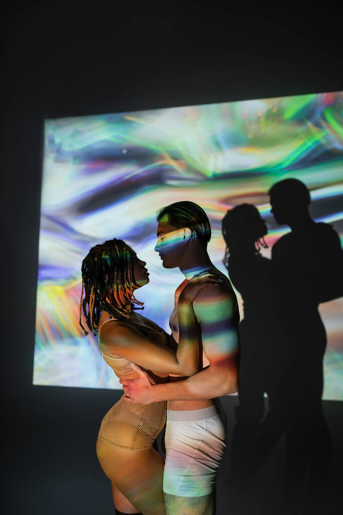 Seitenansicht eines jungen, schulterfreien und muskulösen Mannes, der eine attraktive Afroamerikanerin mit Dreadlocks umarmt, im Netzbody auf schwarzem Hintergrund mit farbenfrohen Projektionen und Lichteffekten - Foto, Bild