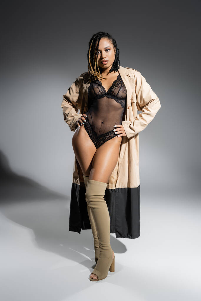 femme afro-américaine pleine longueur avec dreadlocks, en manteau beige à la mode, body sexy en dentelle noire et bottes au-dessus du genou debout avec les mains sur les hanches et en regardant la caméra sur fond gris - Photo, image