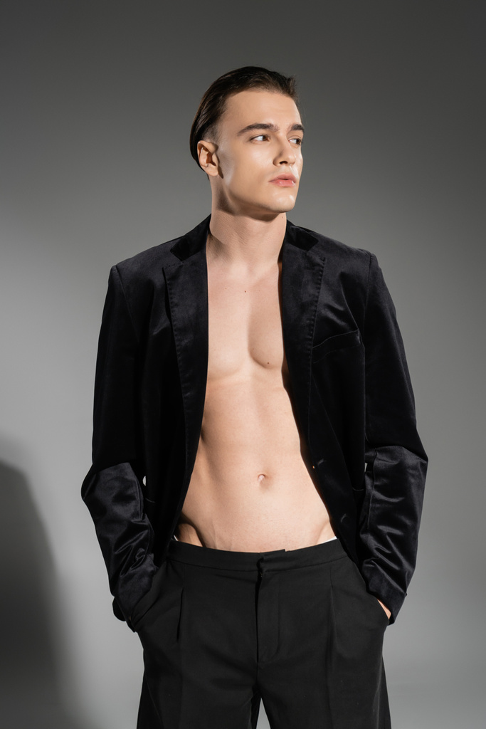 junger, sexy und modischer Mann mit schwarzem Seidenblazer am schulterfreien muskulösen Oberkörper, der wegschaut, während er mit den Händen in Hosentaschen auf grauem Hintergrund posiert - Foto, Bild