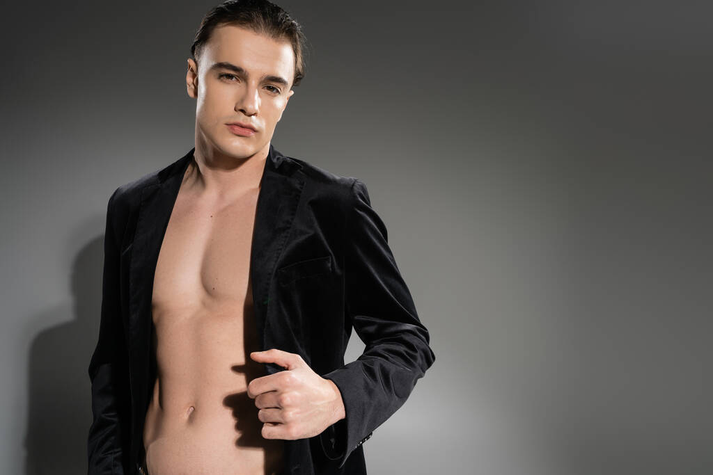 νέος, με αυτοπεποίθηση και όμορφος άντρας με καστανά μαλλιά, φοράει μαύρο και μεταξωτό σακάκι σε γυμνό μυϊκό σώμα ενώ κοιτάζει την κάμερα σε γκρι φόντο - Φωτογραφία, εικόνα