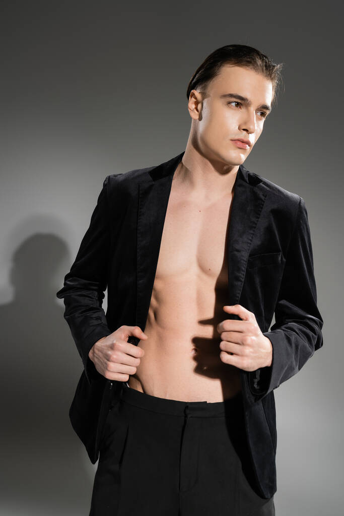 young, self-assured and fashionable man with brunette hair posing in black and silk blazer on shirtless muscular body and looking away on grey background - Photo, Image