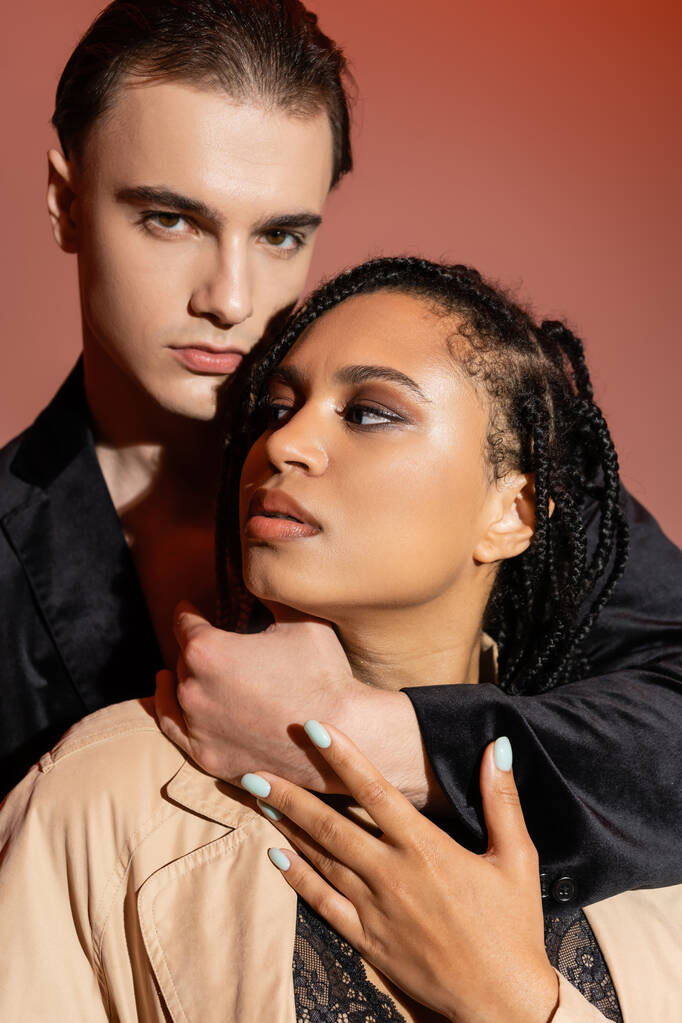 young charismatic man in black silk blazer looking at camera while hugging neck of sexy african american woman with dreadlocks and smokey makeup on pinkish beige background - Photo, Image
