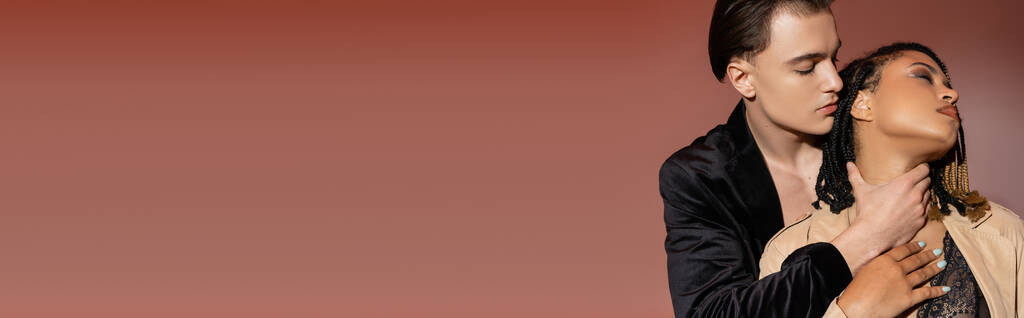μοντέρνος και όμορφος άντρας σε μαύρο μεταξωτό σακάκι αγκαλιάζει το λαιμό της παθιασμένης αφρικανικής αμερικανικής γυναίκας με dreadlocks ντυμένος με δαντελωτά εσώρουχα και καμπαρντίνα σε ροζ μπεζ φόντο, banner - Φωτογραφία, εικόνα