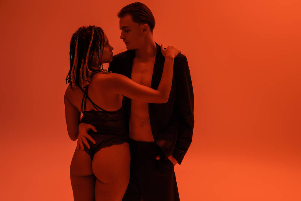 fiatal és elegáns férfi áll kéz a zsebben fekete rövidnadrág és ölelés vonzó afro-amerikai nő szexi fenék, visel csipke test ruha narancssárga háttér piros világítás - Fotó, kép