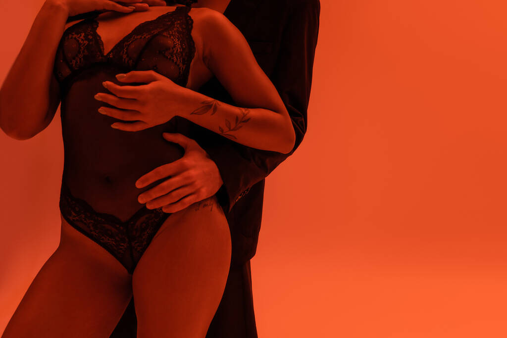 vue partielle du jeune homme en blazer noir embrassant femme américaine africaine sexy et passionnée avec bras tatoué portant un body en dentelle sur fond orange avec effet de lumière - Photo, image