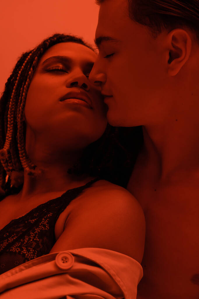 young, shirtless and sexy man seducing and kissing passionate african american woman with dreadlocks and closed eyes, wearing black lace lingerie on orange background with red lighting effect - Photo, Image