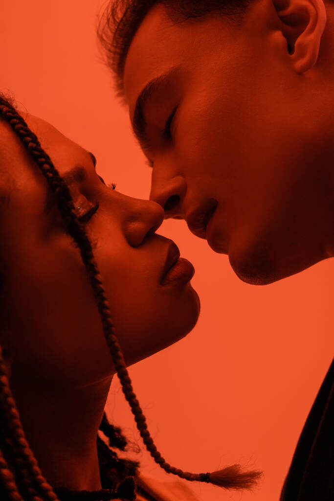 close up view of young and sexy interracial couple in love, young man and african american woman with dreadlocks kissing with closed eyes on orange background with red lighting effect - Photo, Image