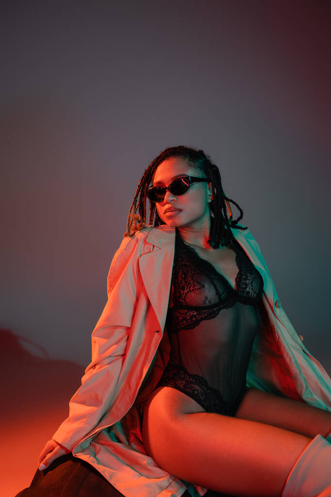 expressive and fashionable african american model in dark sunglasses, black lace bodysuit and beige trench coat sitting on huge tire and looking away on grey background with red lighting - Photo, Image