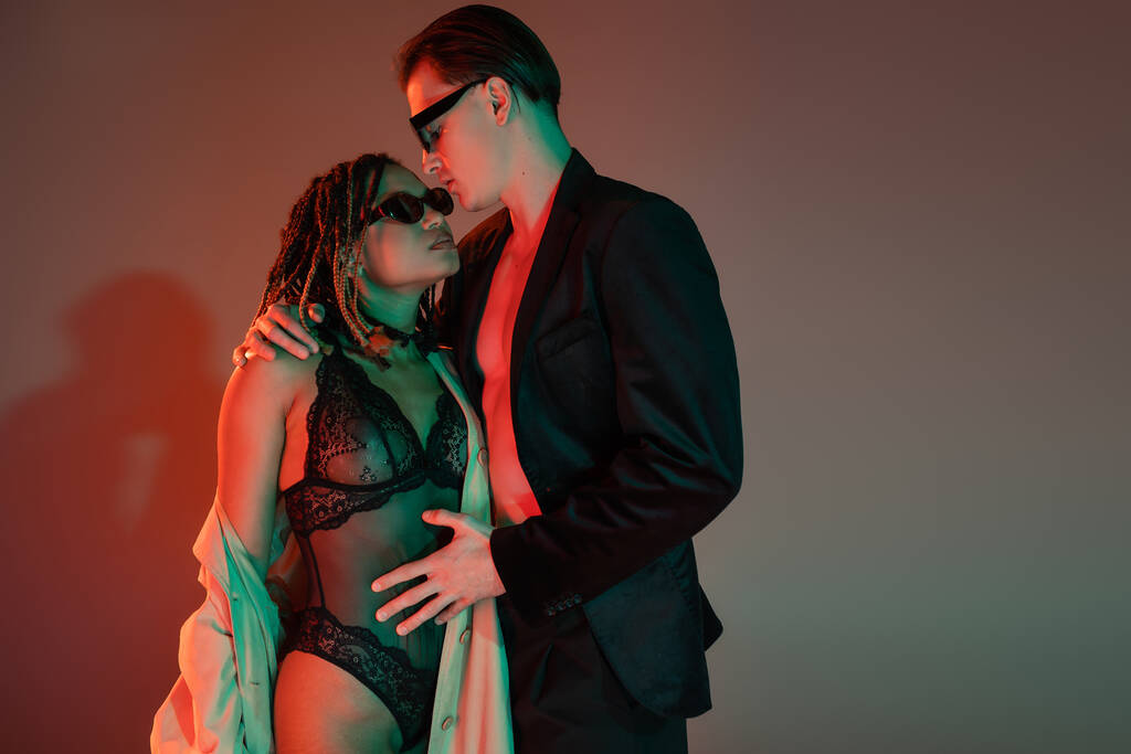glamorous man in dark sunglasses and blazer embracing provocative african american woman in black lace bodysuit and beige trench coat on grey background with red lighting - Photo, Image