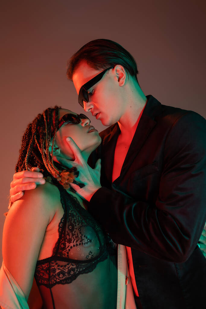 молодий і самовпевнений чоловік в темних сонцезахисних окулярах і чорний блейзер спокушає чарівну афроамериканську жінку в мереживному костюмі на сірому фоні з червоним освітленням
 - Фото, зображення