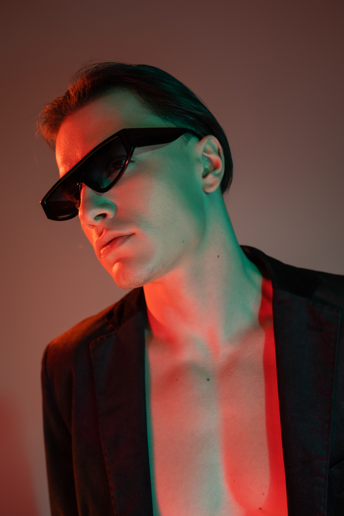 портрет молодого голого чоловіка в чорному модному блістері і темних стильних сонцезахисних окулярах, дивлячись далеко, позуючи на сірому фоні з червоним освітленням
 - Фото, зображення